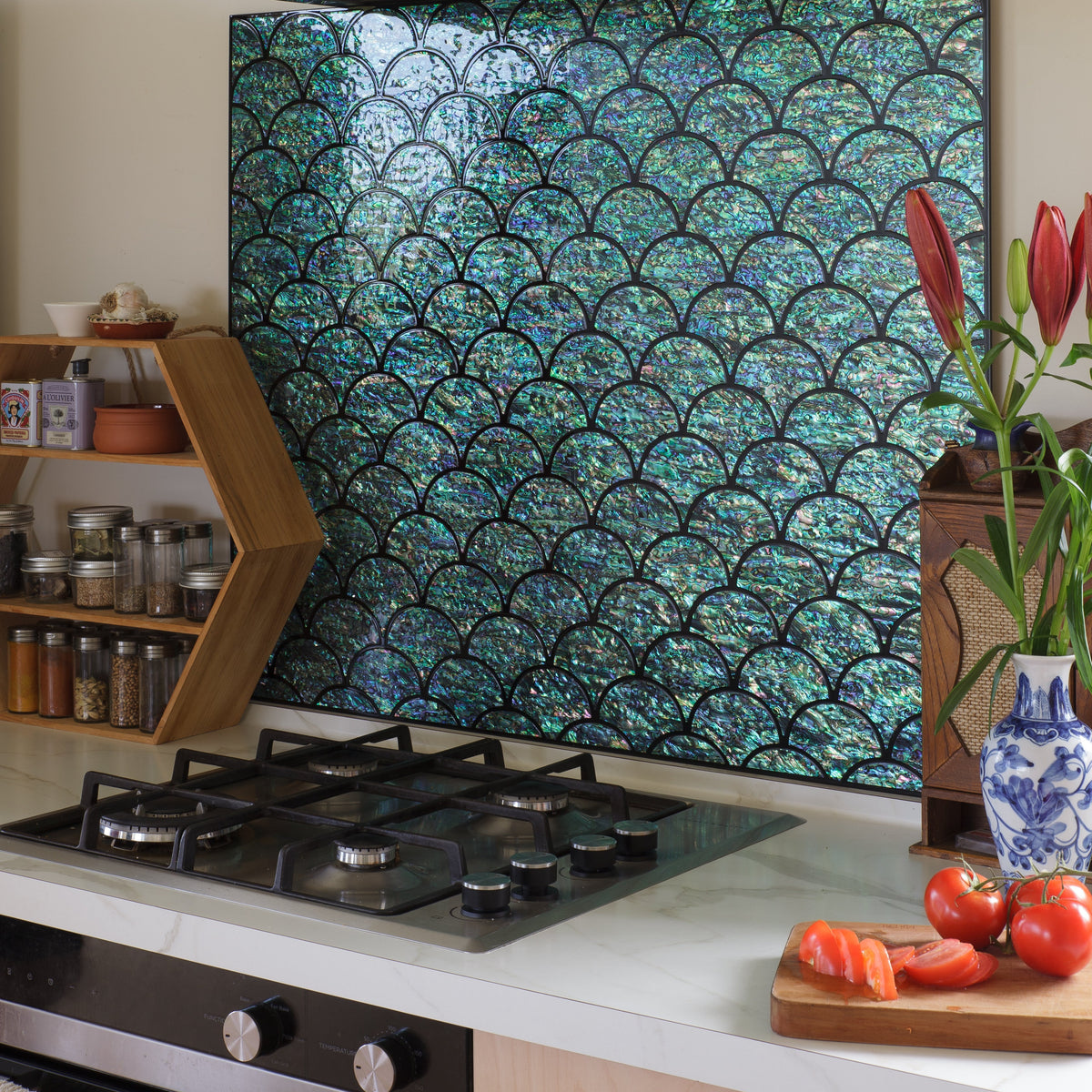 A kitchen backsplash featuring a large rectangular abalone tile panel with a black and teal-green scallop or fish-scale pattern, installed above a gas stovetop on a white countertop.