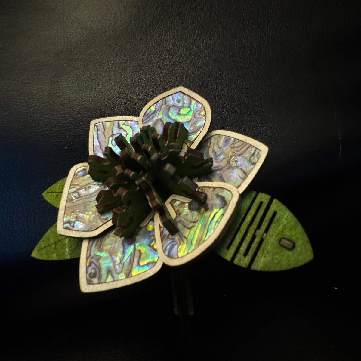A handmade flower-shaped brooch featuring iridescent abalone shell petals with green and blue shifts, a dark green central cluster, and green felt leaves on a black background.
