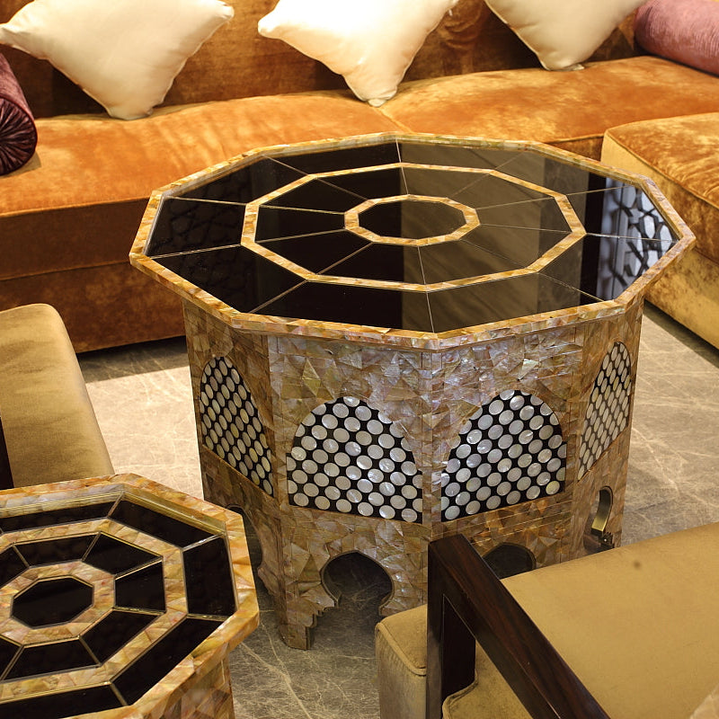 An octagonal mosaic coffee table with a black and gold geometric glass top and a mother-of-pearl shell base, shown in a furnished living room with matching chairs and sofas.