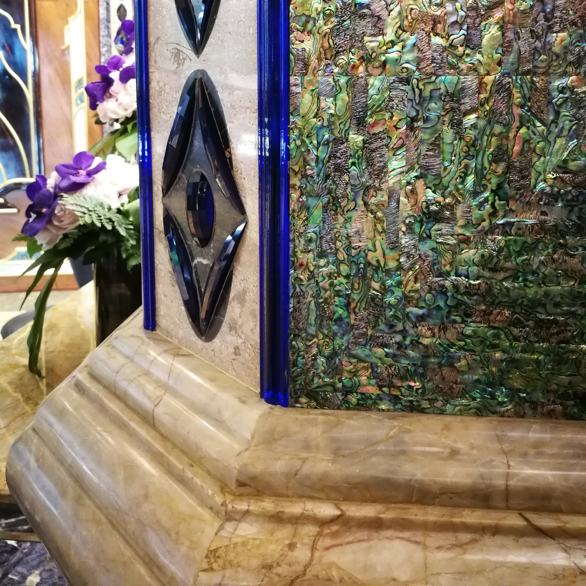 A close-up of an ornate decorative column feature with a beige marble base, blue glass edging, and a textured abalone panel in green, gold, and blue tones. Purple flowers are visible at the left edge.