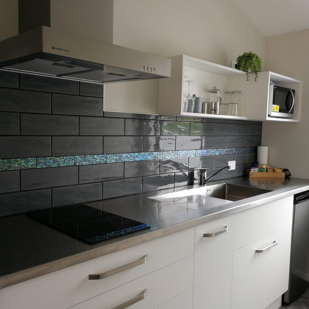 A modern kitchen setup featuring white cabinets, a black tiled backsplash with a blue paua abalone accent stripe, a stainless steel sink and cooktop, and an open shelving unit with glassware and a small potted plant.