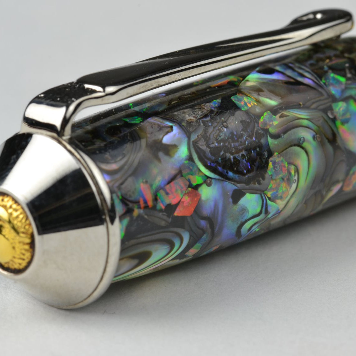 A close-up of a luxury pen featuring a paua abalone shell handle with iridescent green, blue, and purple hues, accented by polished silver-tone metal and gold-tone details.