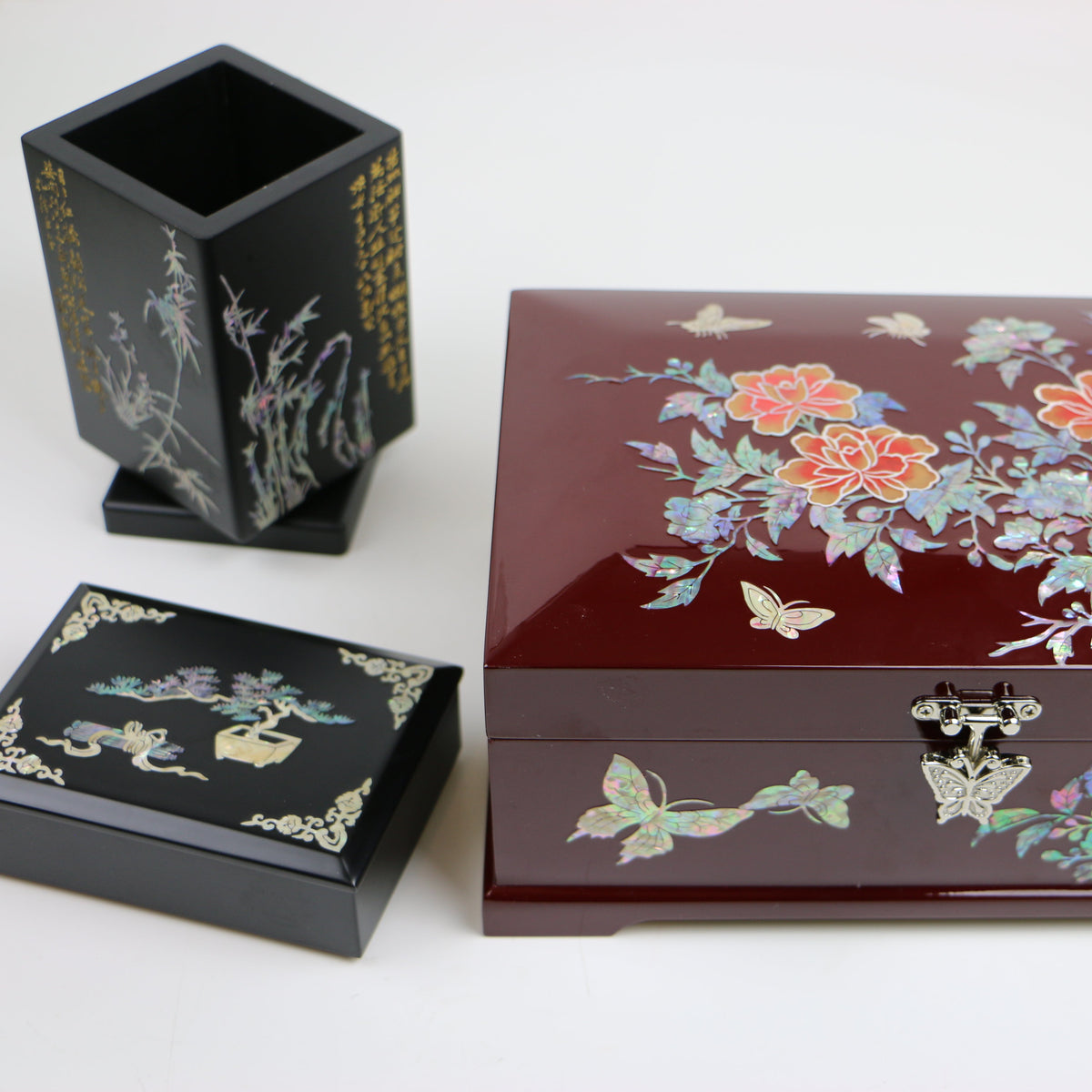 Three decorative lacquer boxes on a white background: a large rectangular reddish-brown box with floral abalone inlay, a smaller square black box with gold detailing, and a tall black hexagonal box with botanical abalone inlay.