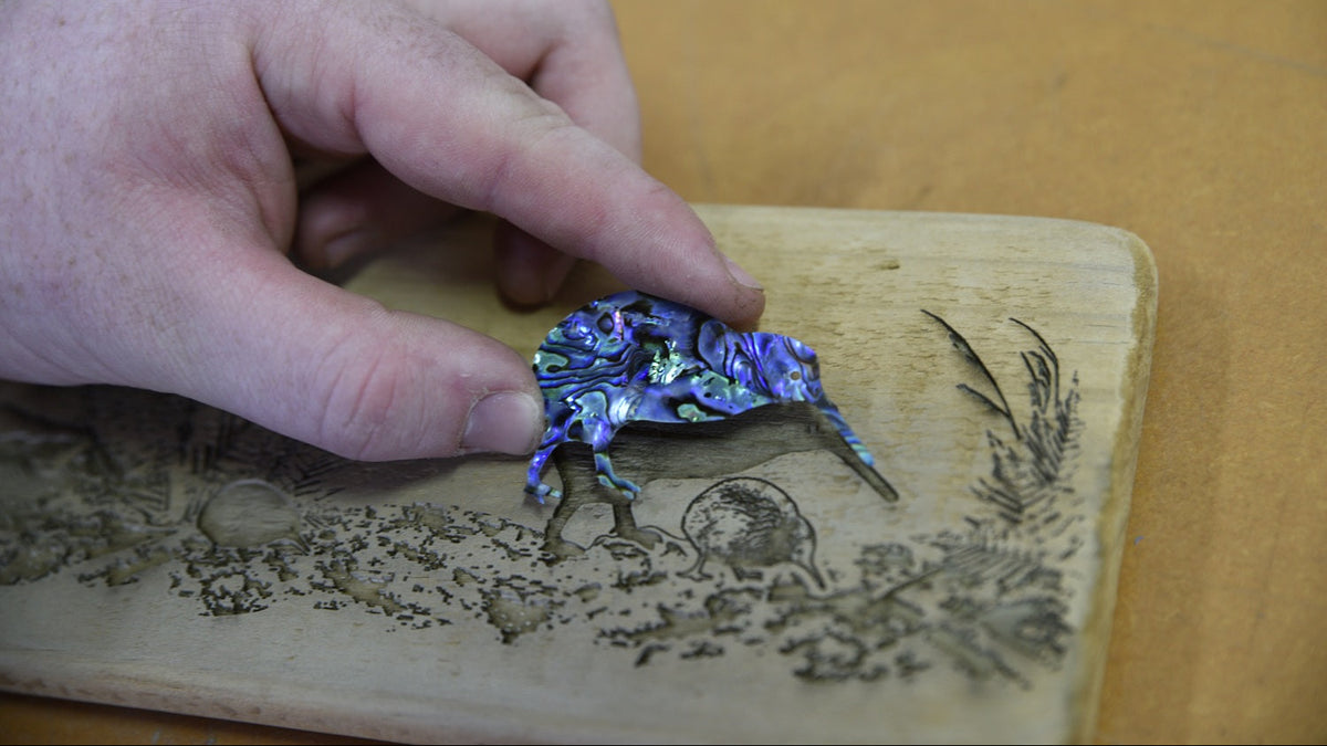 A hand holds a small abalone shell pendant shaped like a kiwi bird over a hand-burned wooden plaque on a tan surface.