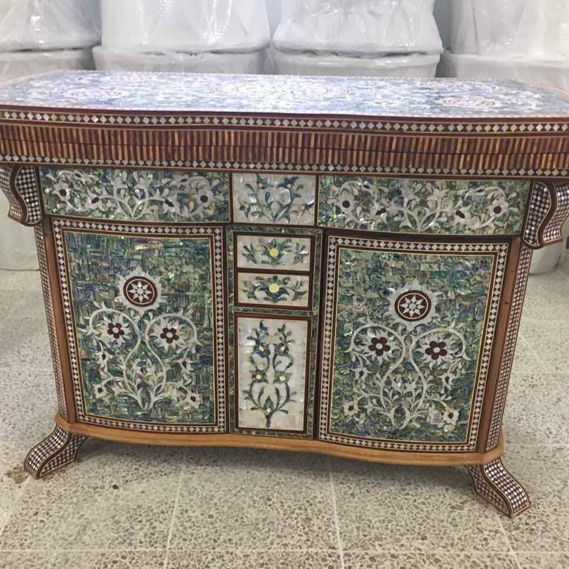 An ornate traditional wooden cabinet decorated with intricate floral shell inlay patterns in green, blue, and white, featuring a mosaic-style top and carved legs, photographed in a showroom.